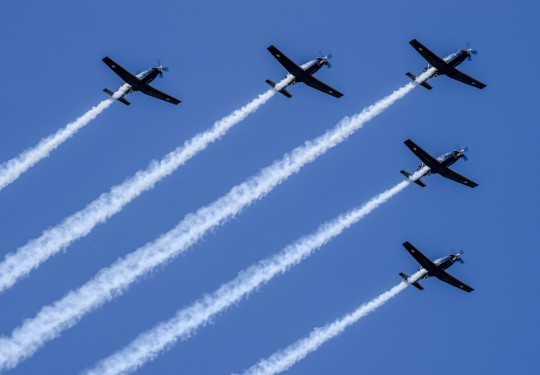 A formation flight of five aircraft in the shape of an arrowhead soars in the blue sky with white trails behind.
