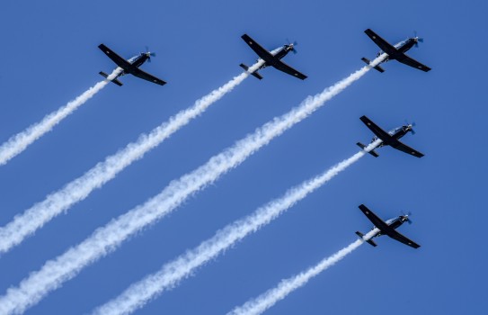 A formation flight of five aircraft in the shape of an arrowhead soars in the blue sky with white trails behind.