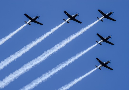A formation flight of five aircraft in the shape of an arrowhead soars in the blue sky with white trails behind.