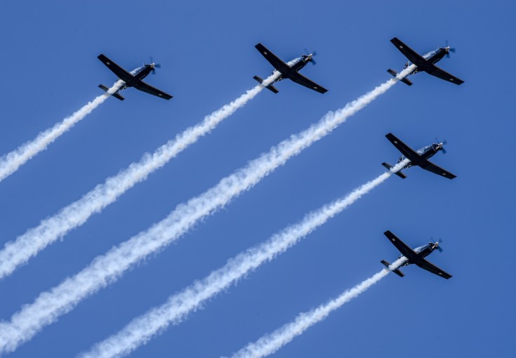 A formation flight of five aircraft in the shape of an arrowhead soars in the blue sky with white trails behind.