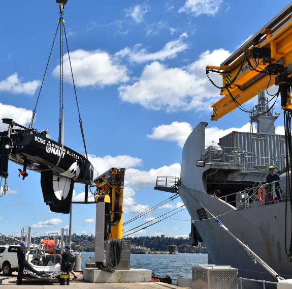 A small black USV is craned onto a large grey ship at port in front of blue skies and scattered clouds.