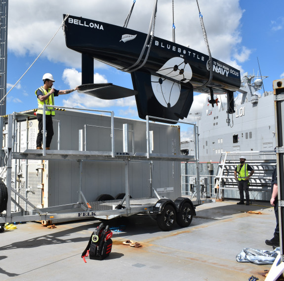 A small black USV is craned onto a large grey ship as crew in high visibility vests and helmets guide it into position.