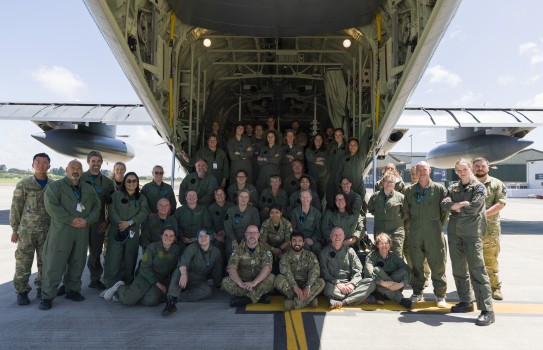 A group poses at the tail of a large aircraft, the ramp is open and people stand on top as well as on both sides of the aircraft.  There are four rows of males and females, they all wear military flying suits and a variety of footwear as they pose for the