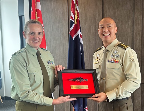 Two people in army uniforms hold a frame in front of the flags of Singapore and New Zealand.