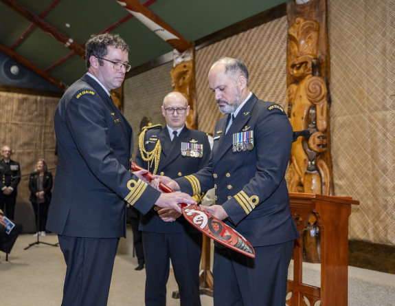 Three uniformed stand together. Two of them are holding a carved hoe (paddle) - a handover. They are in a marae and there are others behind them in the background. 