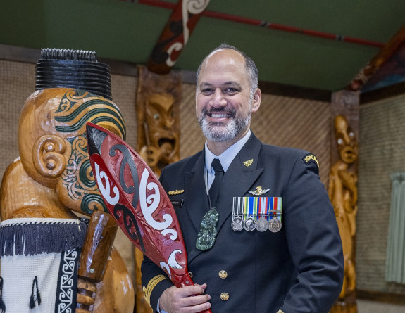 A uniformed Navy sailor stands smiling, looking at the camera holding a carved hoe (paddle) in a marae. 