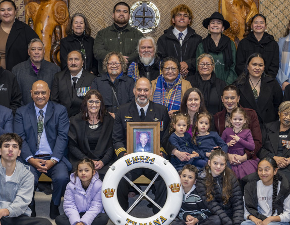 A uniformed sailor sits in the middle of a large group of people. The people are all looking at the camera. There is a Navy life ring with HMNZS Te Mana written on it and the sailor is holding a picture frame with a photo in it. 