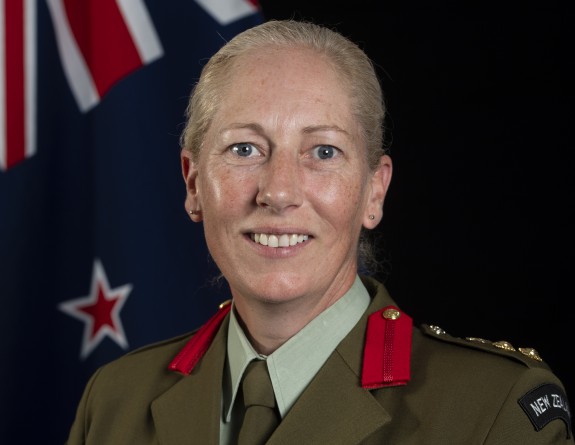 A woman wearing an Army uniform with medals on her jacket smiles at the camera in front of a New Zealand flag. 