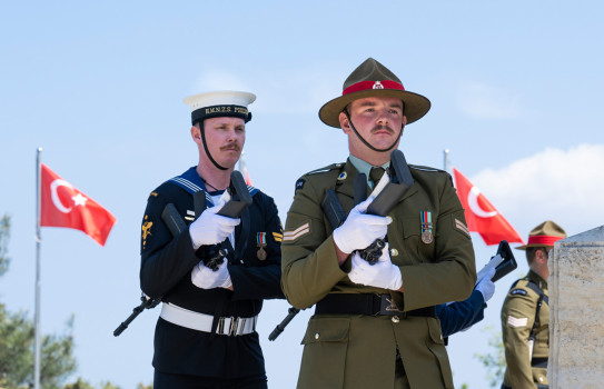 A sailor and soldier in full military dress stand in front of blue skies with Türkiye flags blowing in the background.