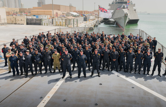 A large group of personnel stand with their arms behind their backs, legs shoulder-width apart on the flight deck of HMNZS Te Kaha.