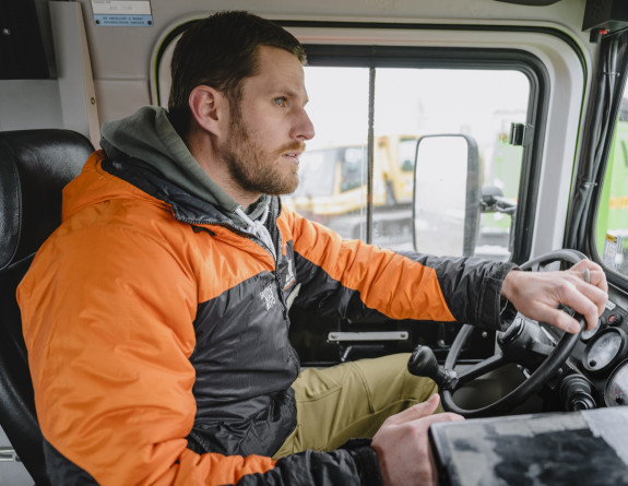 Cam driving a large vehicle while wearing an orange and black Antarctica NZ jacket.