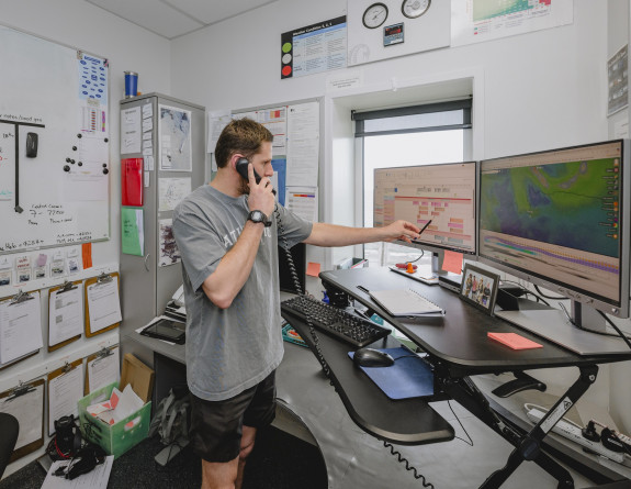 Cam in t-shirt and shorts in an office, pointing at a screen while on the phone.