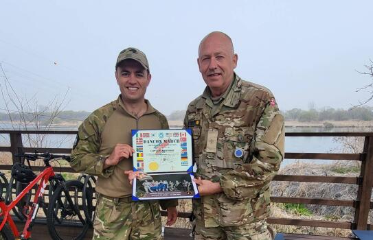 Two military personnel in uniform standing in uniform holding a certificate. In the background is some water, behind a fence. There's also a red bike to the left of the photo. 