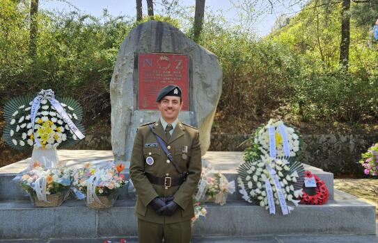 A solider stands in uniform, in front of a memorial (stone like monument). There are wreaths on the memorial. 