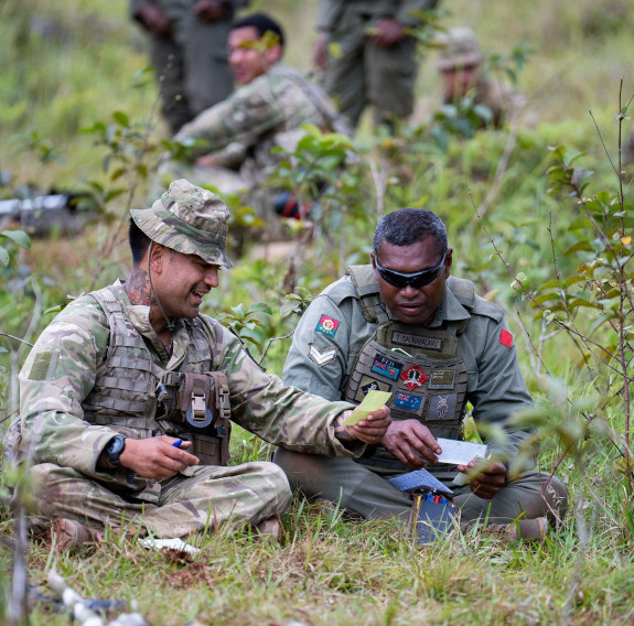 A soldier from the New Zealand Army (left) and one from the Republic of Fiji Military Forces (right) sit on the grass during a discussion during a break in the action. Other soldiers are in the background.