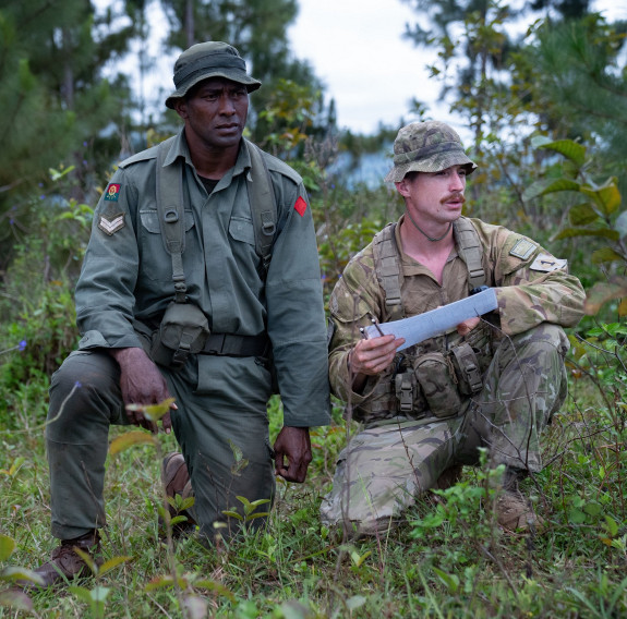 A soldier from the Republic of Fiji Military Forces (left) takes a knee while looking in the distance. A New Zealand Army soldier holds a document while also taking a knee in front of the trees of the jungle.
