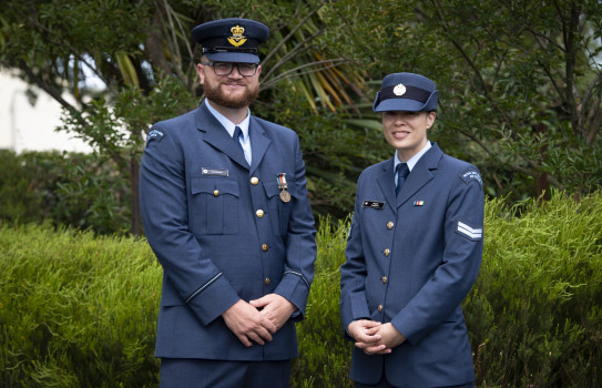 Two airman stand looking at the camera, side-by-side in their uniform. They hands are clasped together in front of each of them.