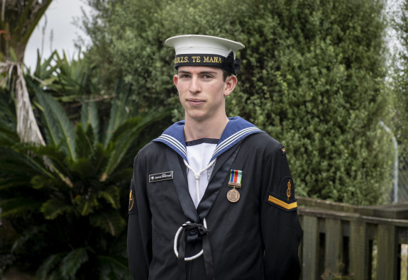 Portrait of a Navy sailor wearing formal navy uniform with head dress. He's standing outside infront of some nice greenery. 