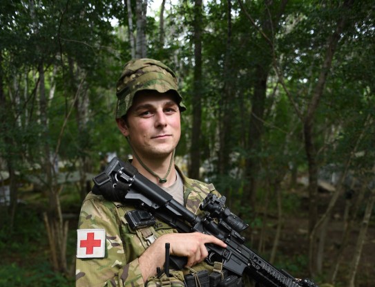 A soldier stands in uniform holding a weapon, staring at the camera and in the bush. 