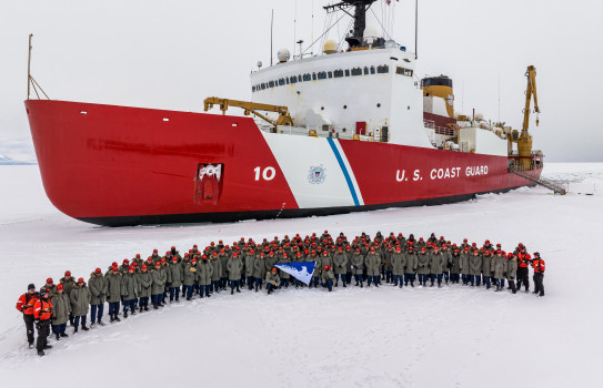 150 crew wearing green jackets and red baseball caps stand on the ice with the USCGC Polar Star stationary behind them.