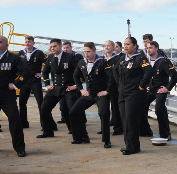 A group Navy sailors stand with hands on their hips as they perform a haka, a cultural dance on the wharf.
