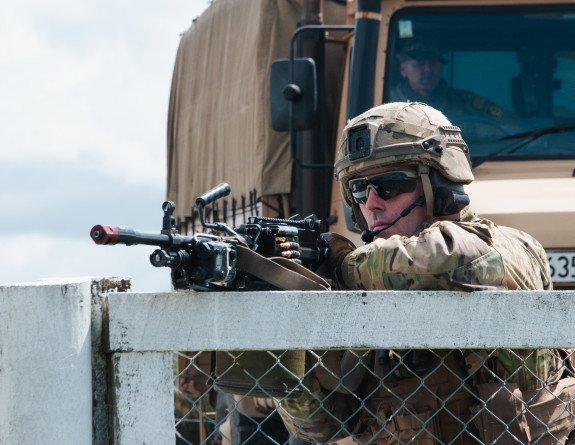 A soldier aims their weapon on the top of a white wooden framed, chain fence with a tan Army vehicle behind them.