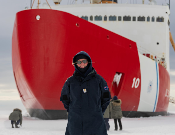MID Walker in front of the USCGC Polar Star on the ice. She is wearing navy blue jacket and pants, sunglasses and a big smile.