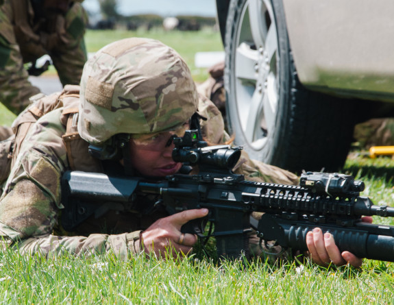 A soldier looks into the scope of their rifle while laying on the grass in front of a vehicle.