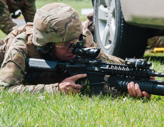 A soldier laying on the grass alongside a vehicle, aiming through the sights of their weapon.