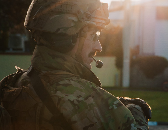 A soldier wearing a camouflage uniform, helmet and glasses stands guard as the sun flares above them.