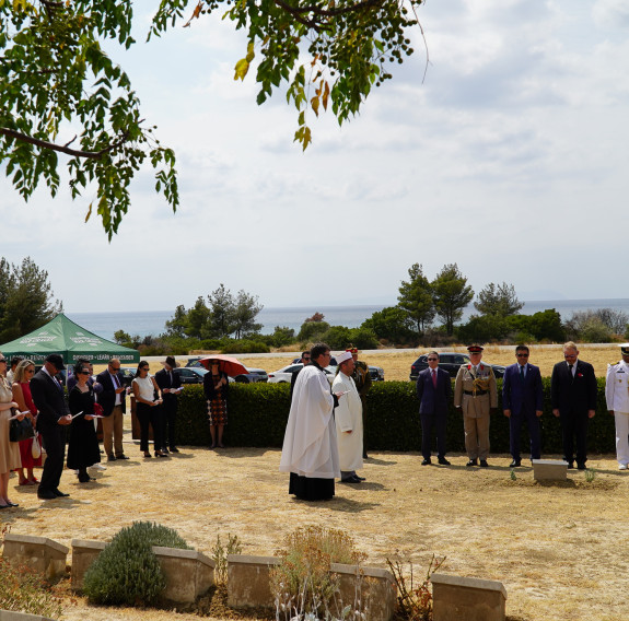 A service takes place next to graves in a field of dry grass, under a tree.