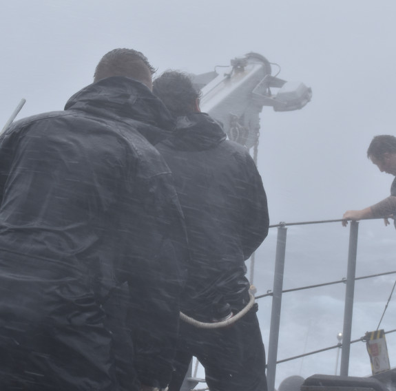 Three sailors stand on the flight deck of HMNZS Te Mana in the wind and rain, the sailors are drenched with water and visibility is extremely poor.