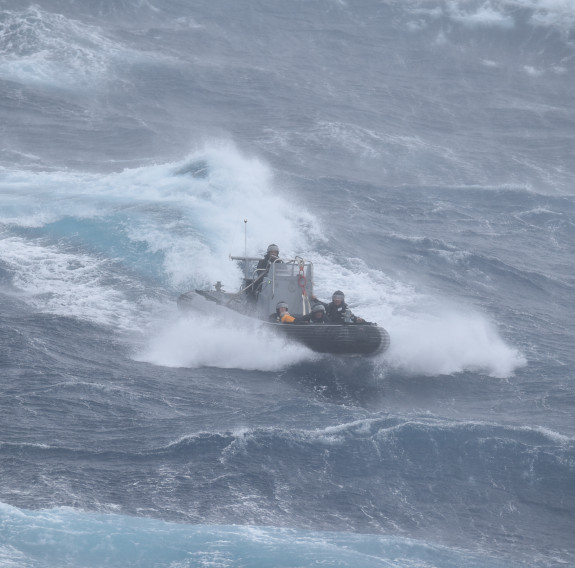 A RHIB cuts through the top of a wave during the rescue, the wind blows the water off the front of the vessel. 