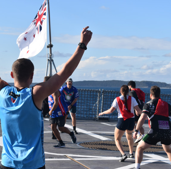Sailors play sports on the flight deck. In the foreground a sailor wearing a blue singlet lifts his arms as others are focussed on the game in the background.
