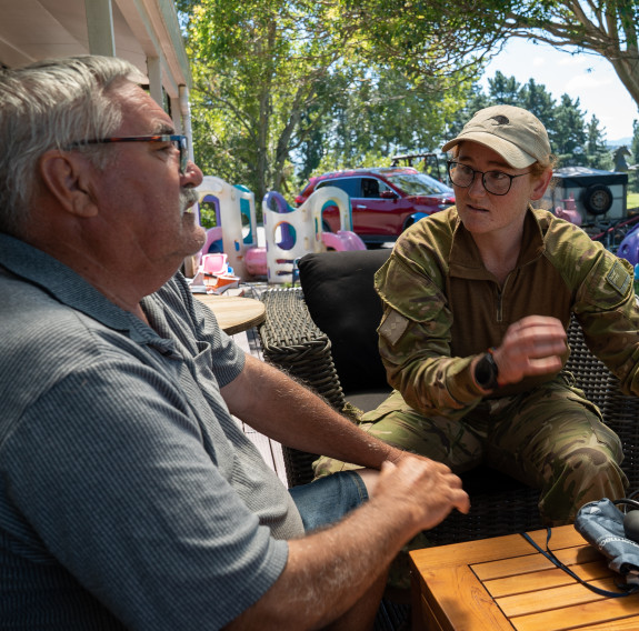 A man with grey hair (left) wearing a grey shirt sits on the front deck outside a house while talking to a New Zealand Army medic (right) wearing camouflage uniform a beige cap and glasses. In the background there is green lawn in front of a hedge and tre