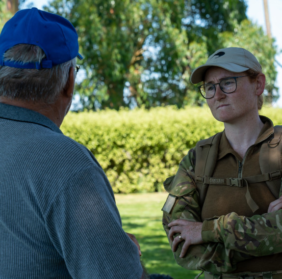 A man facing away (left) with grey hair, a grey shirt and blue hat talks to a New Zealand Army medic (right) wearing camouflage uniform with a red cross on her left arm, a beige cap and glasses. They stand on a green lawn in front of a hedge and trees.