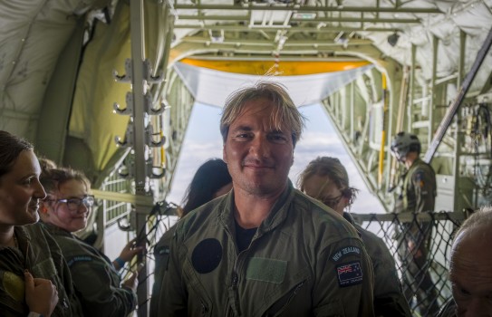 A blonde haired male looks into the camera, he is wearing a green military flying suit.  Behind him are several others wearing the same flying suit. As the aircraft flies, the back door is open and views can be partially seen of the clouds behind.