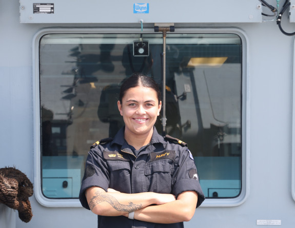 Sub Lieutenant Emily Aull smiles as she stands with her arms crossed, underneath the HMS Spey ship badge. She is wearing her Navy blue uniform with black and gold accents. 