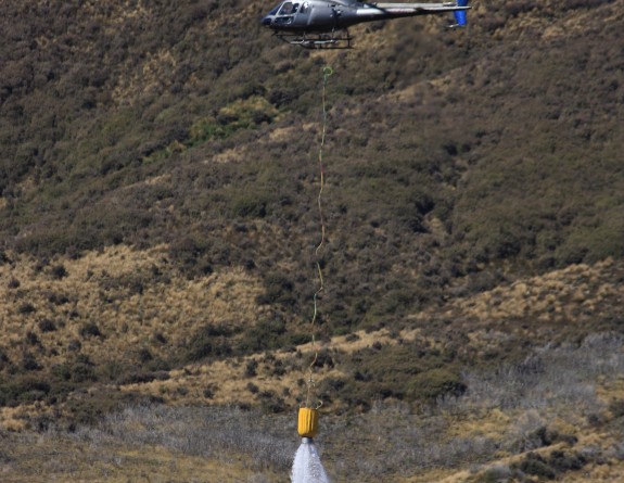 A single helicopter flies through brown hilly countryside releasing water from the bucket it is carrying.