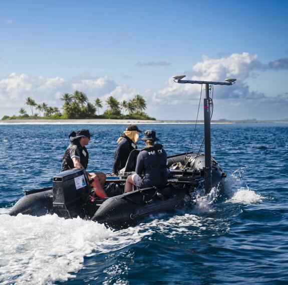 Three people in a small boat at sea conduct surveying with equipment attached to the side of the boat. In the background is a small tropical island.
