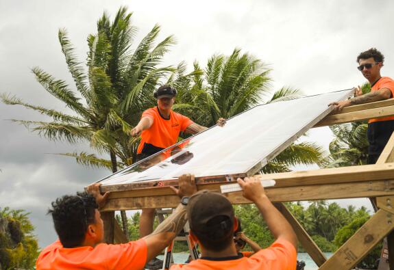 Four soldiers in orange high vis shirts life a solar panel onto a frame in front of a palm tree