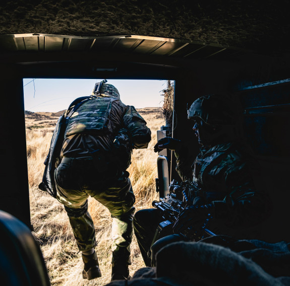A uniformed soldier steps out of a vehicle that is dark inside and steps out to the sunlight. There is another soldier still in the vehile.