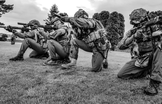 The image is black and white. Four soldiers on one knee, on the grass on the grass in full uniform, holding weapons. None are looking at the camera, three are directing their weapon to the left of the photo and one to the right.