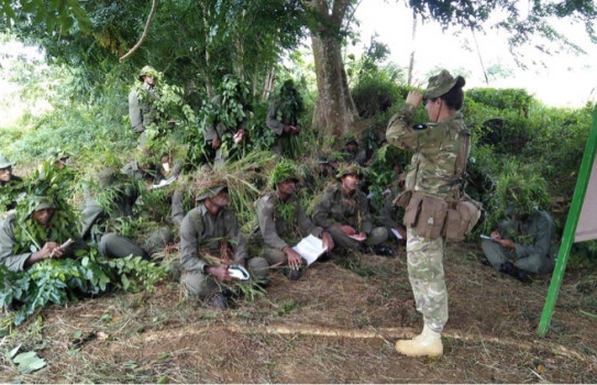 CPL de Schot delivers a lesson reviewing cam and concealment in the jungle environment. Soldiers are sitting on a mound under a tree in camouflage listening to the Corporal.