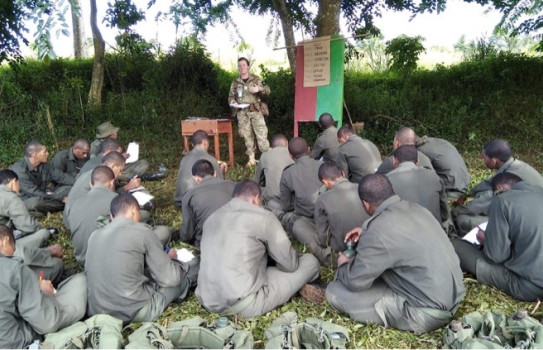 CPL de Schot delivers a lesson on the cycling process of the M16 Rifle. The soldiers sit on the ground in the dense bush and take notes.