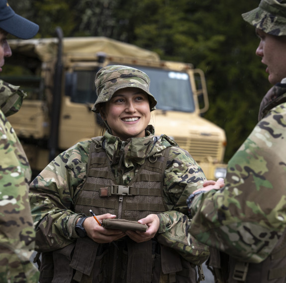 Three aviators talk in front of a Unimog (a large tan-coloured military vehicle)