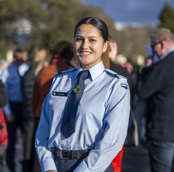 An aviator stands in front of a crowd of people smiling at the camera as they sun shines on their skin. 