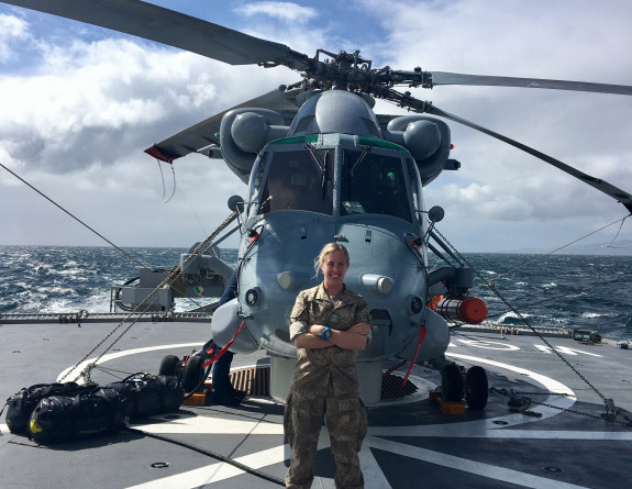 A nurse stands in front of a Seasprite helicopter with arms crossed on the flight deck of a ship. Scattered clouds, blue sky and white small white capped waves of the ocean in the background.