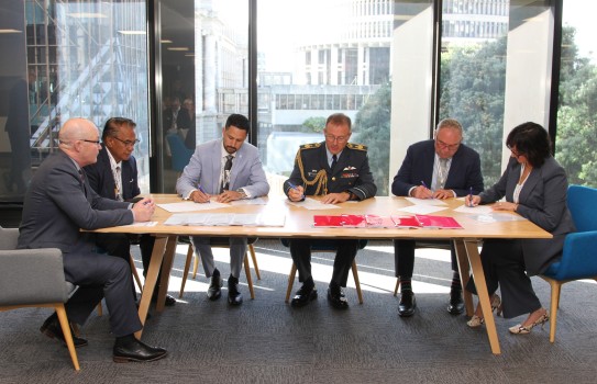 Various people including a military dressed officer sit at a table signing contracts. Behind them through the glass window is the Beehive building.