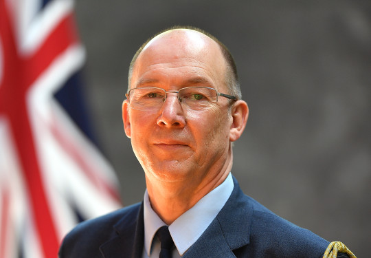 Wing Commander Graham Streatfield in full military dress uniform, standing in front of the New Zealand flag smiling.